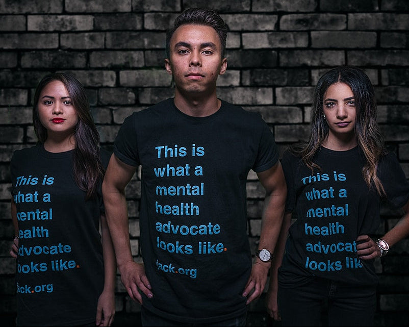 Three people wearing black t-shirts with a message about mental health advocacy against a brick wall.