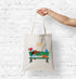 Tote bag with a design of a dog and birds on a bench held by a hand against a white brick wall.