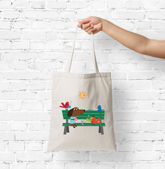 Tote bag with a design of a dog and birds on a bench held by a hand against a white brick wall.